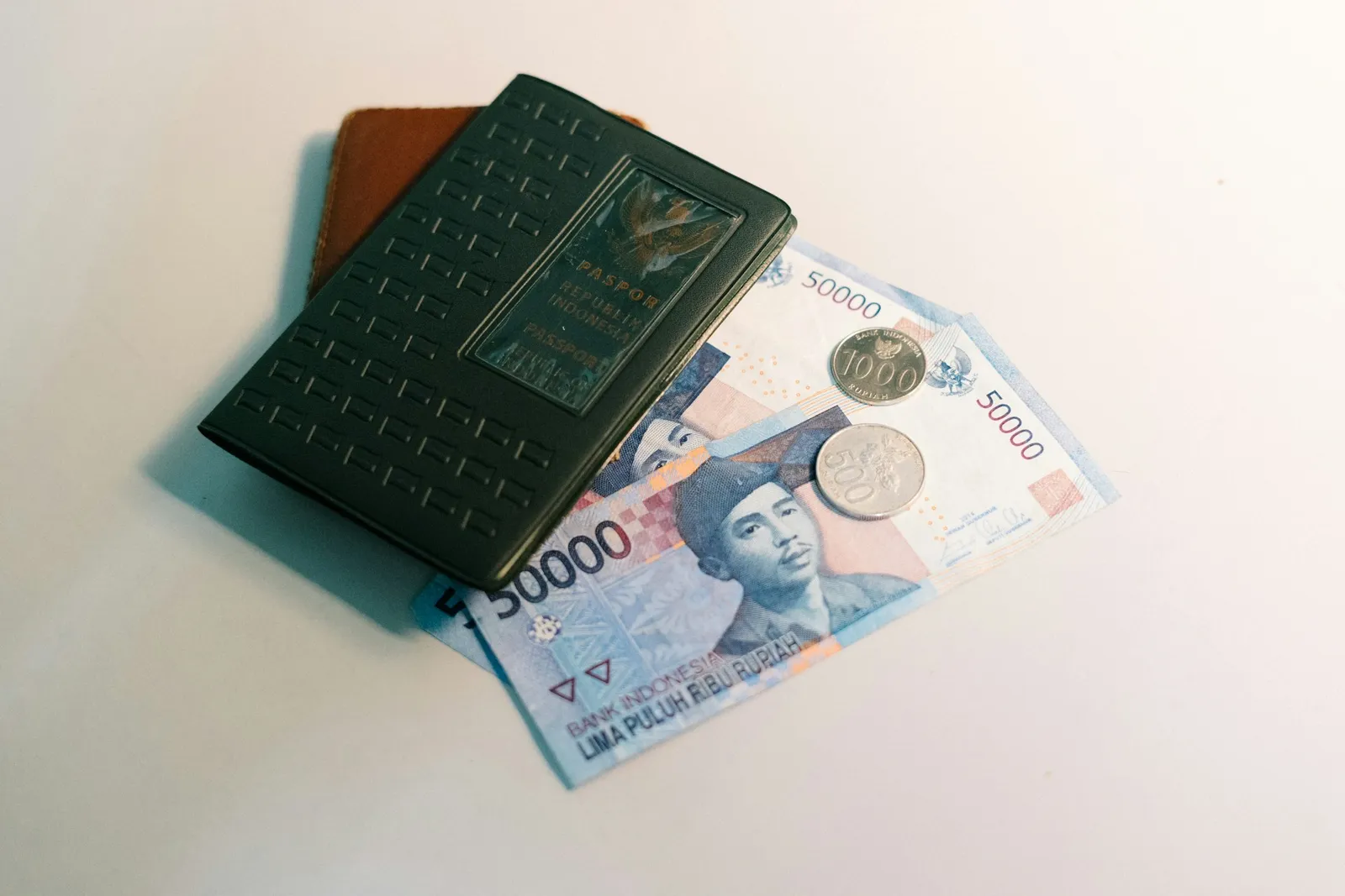 Flat-lay of a passport, a brown leather wallet, and Indonesian rupiah banknotes arranged on a clean white surface
