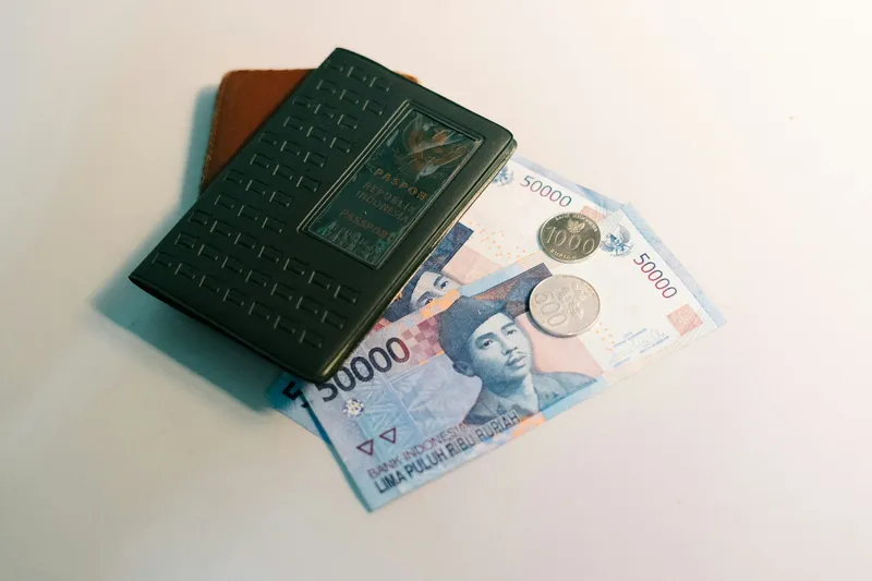 Flat-lay of a passport, a brown leather wallet, and Indonesian rupiah banknotes arranged on a clean white surface