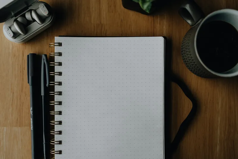 Flat-lay of an open notebook, ballpoint pen, headphones, and a cup of coffee arranged on a wooden desk
