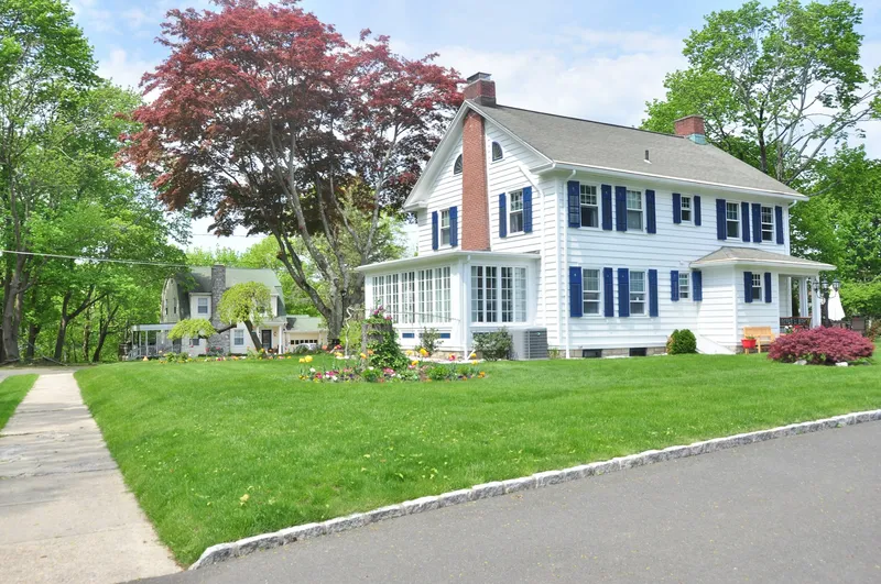 White and gray two-story colonial-style home with green front lawn and mature trees in the background under a clear daytime sky