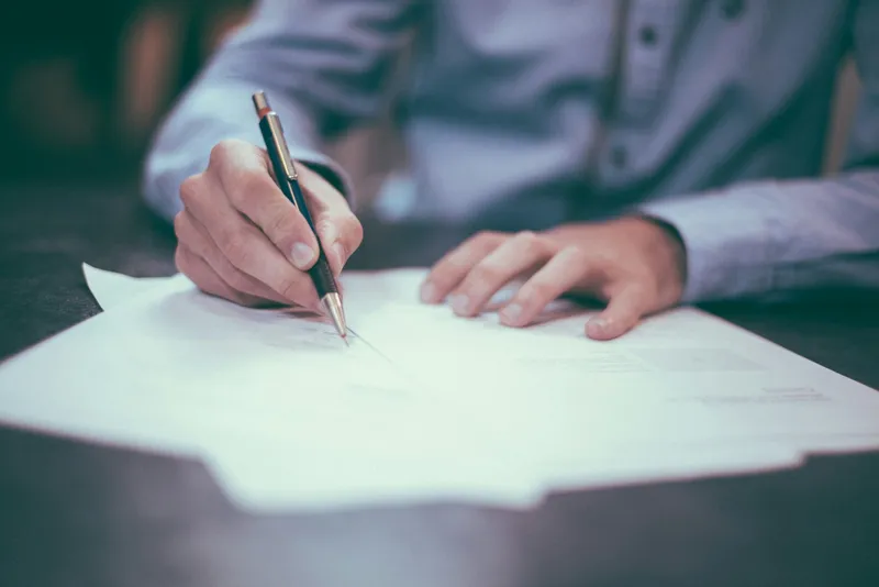 Close-up of a man's hand writing on a paper document at an office desk, with a soft-focus blurred background