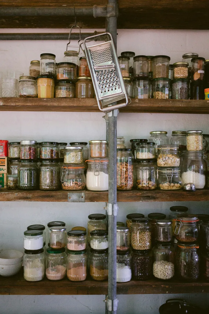 Assorted clear glass jars holding pantry staples lined up on a white wooden shelf with a small cheese grater off to one side