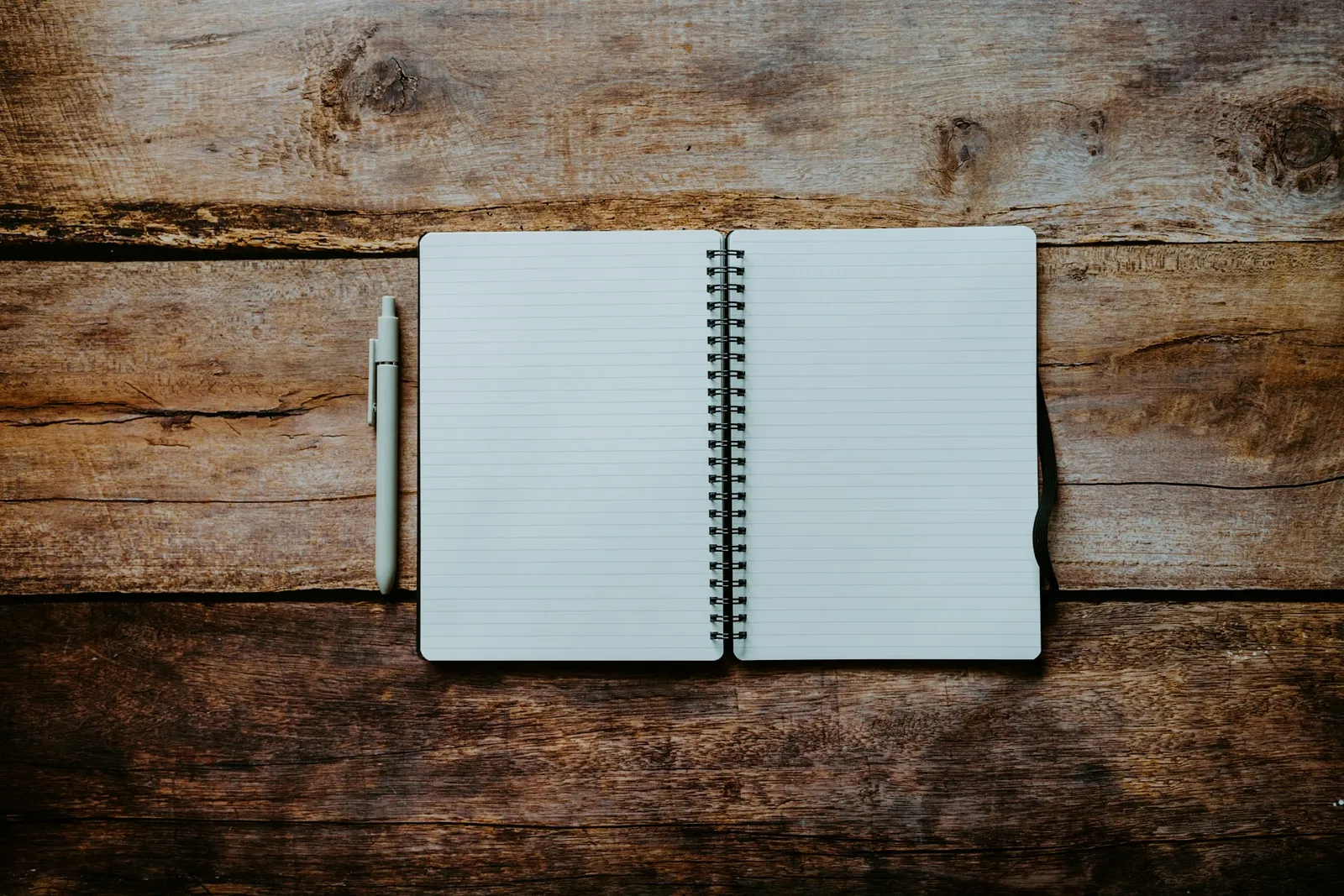 Open spiral-bound notebook with blank lined pages on a raw wooden table next to a pen, top-down view