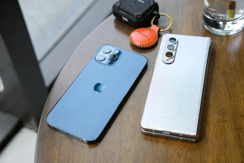 Two smartphones lying face-up on a wooden surface next to a small leather-cased tag, screens dark, photographed from above