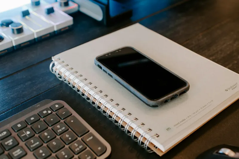 Black smartphone resting face-down on an open spiral notebook beside a dark computer keyboard in a minimalist flat-lay workspace