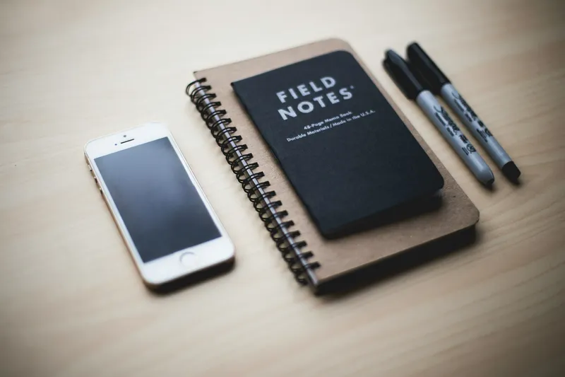 Silver iPhone face-down on a wooden desk next to a Field Notes notebook and a pen